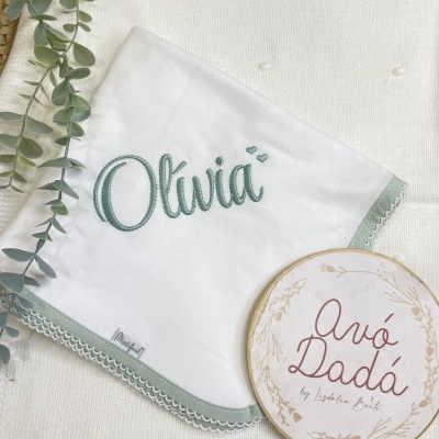 Manta branca bordada com nome Olívia e detalhe verde claro com placa redonda decorativa ao lado