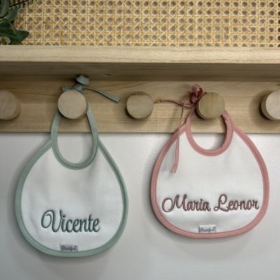 Dois babetes de bebé com nomes bordados pendurados em ganchos de madeira
