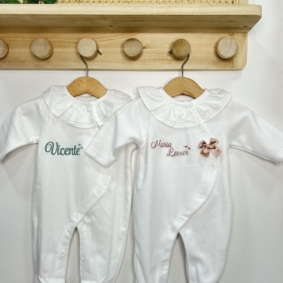 Dois macacões brancos para bebé com nomes bordados pendurados em cabides de madeira num cabide de parede