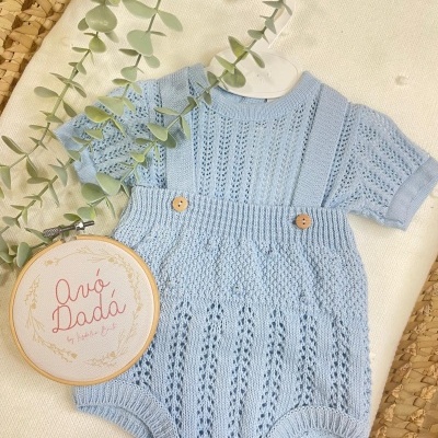 Conjunto de roupa de bebé azul clara em malha com padrão, com alças e botões de madeira sobre tecido claro e plantas decorativas.