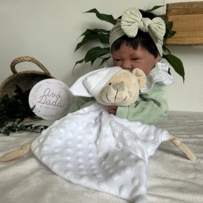 Boneca bebé com laço bege segurando cobertor branco com cabeça de urso de peluche sentada em superfície clara com planta e cesta ao fundo