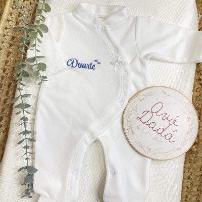 Macacão branco para bebé com bordado azul e laço branco dentro de cesto de palha com manta branca e folhas verdes
