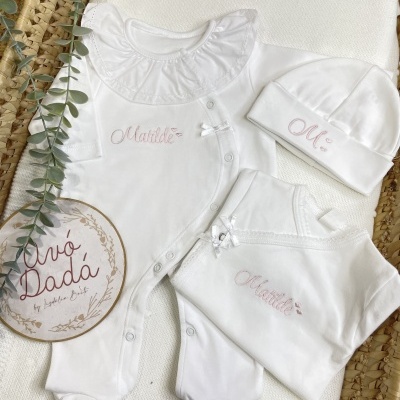 Conjunto de roupa branco de bebé com bordados cor-de-rosa Matilde e M:, com folho e laços, exposto em almofada e cesta de palha.