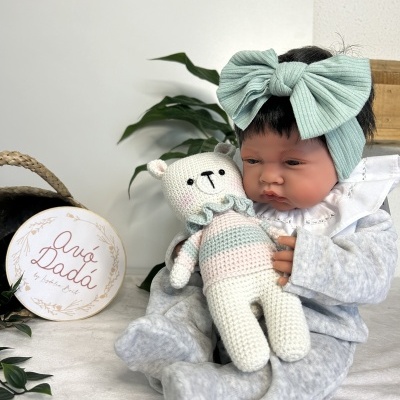 Boneca bebé realista com fato de pijama cinzento e fita de cabelo verde água segurando urso de tricot