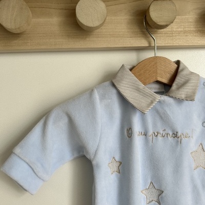 Pelele azul claro com gola riscada bege e branco e estrelas bordadas pendurado em cabide de madeira