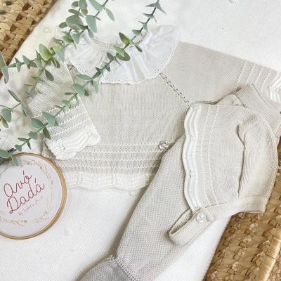 Conjunto de roupa de bebé bege e branco em malha com gola e botões, sobre tapete de palha e fundo branco