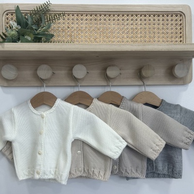 Quatro casacos infantis de malha em tons neutros pendurados em cabides de madeira sob prateleira em madeira com detalhe em palha.