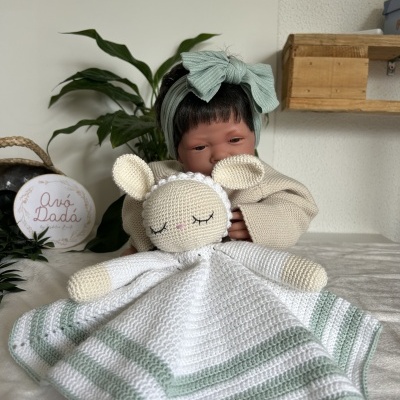 Boneca bebé a segurar cobertor de crochê com ovelha, em cenário com planta e prateleira de madeira