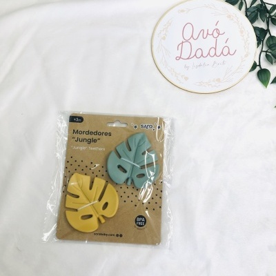 Dois mordedores em forma de folha amarelo e verde em embalagem, sobre tecido branco com raminho verde e círculo decorativo com texto Avó Dadá.