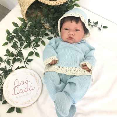 Boneca realista vestida com roupa azul clara e capuz, com placa branca e cesta de palha ao lado.