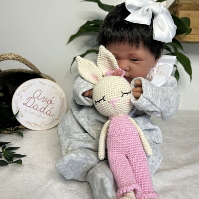 Boneca bebé com fato cinzento e laço branco, segurando coelho de croché rosa e bege