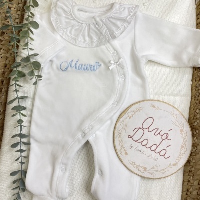 Macacão branco para bebé com gola franzida e bordado azul Mauro, em tecido branco e cesto de palha com planta verde e placa redonda