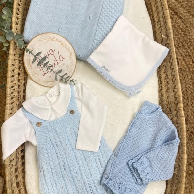Conjunto de roupa azul clara de malha para bebé com body branco e acessórios numa cesta de verga.