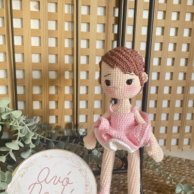 Boneca de croché com vestido rosa sentada com placa branca e planta ao fundo.