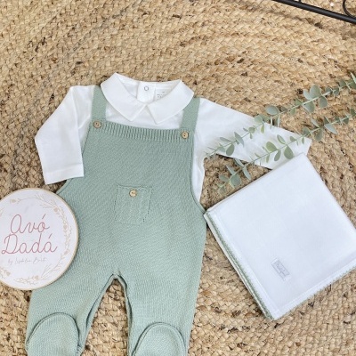 Macacão de bebé verde com camisa branca e placa redonda sobre tapete de fibras.