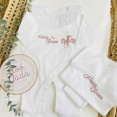 Conjunto de roupa de bebé branco com bordado rosa Maria Teresa e toalha, sobre colchão branco e cesta de vime.