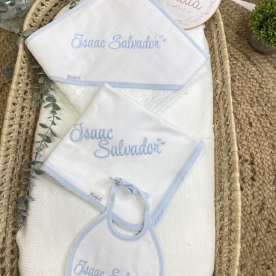 Conjunto de acessórios para bebé brancos e azuis claros com nome bordado, numa cesta de vime