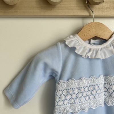 Vestido azul claro infantil com gola branca e renda pendurado em cabide de madeira.