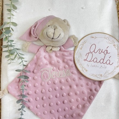 Peluche rosa com textura e nome bordado Teresa sobre tecido branco com malha, ramo verde e placa redonda com texto.