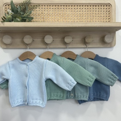 Quatro casacos de malha para bebé em cores pastel pendurados em cabides sob uma prateleira de madeira clara com detalhes em palhinha