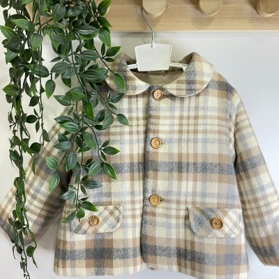 Casaco infantil xadrez bege com botões de madeira pendurado em cabideiro de madeira ao lado de planta verde
