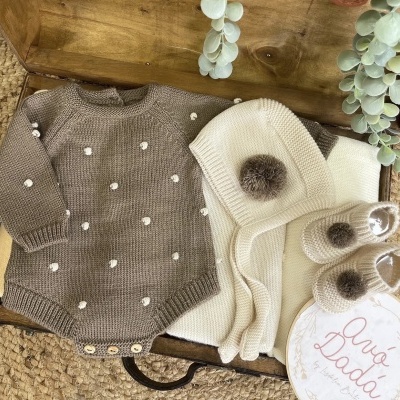 Conjunto de roupa de bebé em malha castanha com bolinhas brancas, touca e sapatinhos bege com pompons castanhos numa mala de madeira.