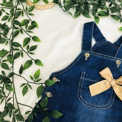 Macacão de ganga azul com alças cruzadas, laço bege e bolso, rodeado por folhas verdes
