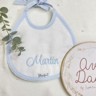 Babete branco com borda azul clara e nome Martin bordado, junto a ramo verde e placa de madeira sobre fundo creme