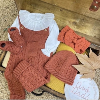Roupa de bebé em malha terracota, camiseta branca e acessórios sobre caixa de madeira
