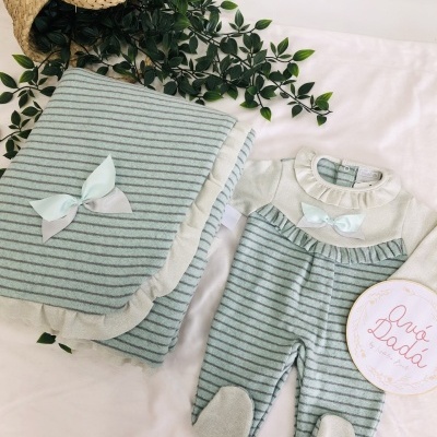 Cobertor e pijama de bebé com riscas e laços brancos, sobre superfície branca, com planta falsa e cesta de palha