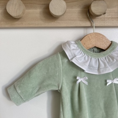 Roupa de bebé verde com gola branca de babados e dois laços brancos pendurada num cabide de madeira