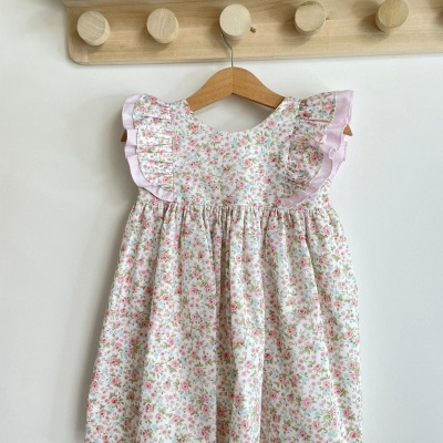 Vestido infantil floral branco com folhos lilases pendurado em cabide numa parede