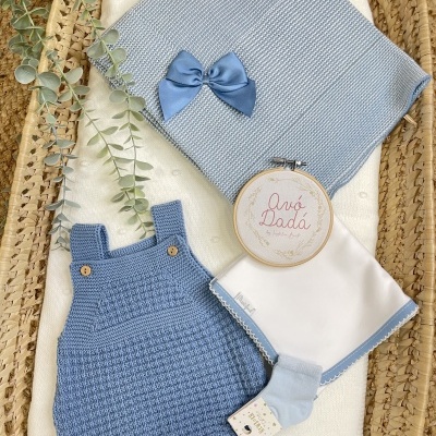 Conjunto de roupas e acessórios para bebé em azul e branco sobre uma cesta de vime.