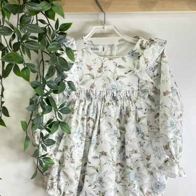 Body para bebé com padrão floral em tons suaves, pendurado num cabide branco junto a uma planta verde e suporte de madeira