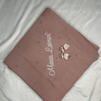 Manta rosa com laço bege e texto bordado 'Manta Lenore' sobre superfície branca