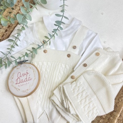 Conjunto de roupa de bebé creme com body branco, jardineira e casaco tricotados, planta verde e placa decorativa com texto Avó Dadá.