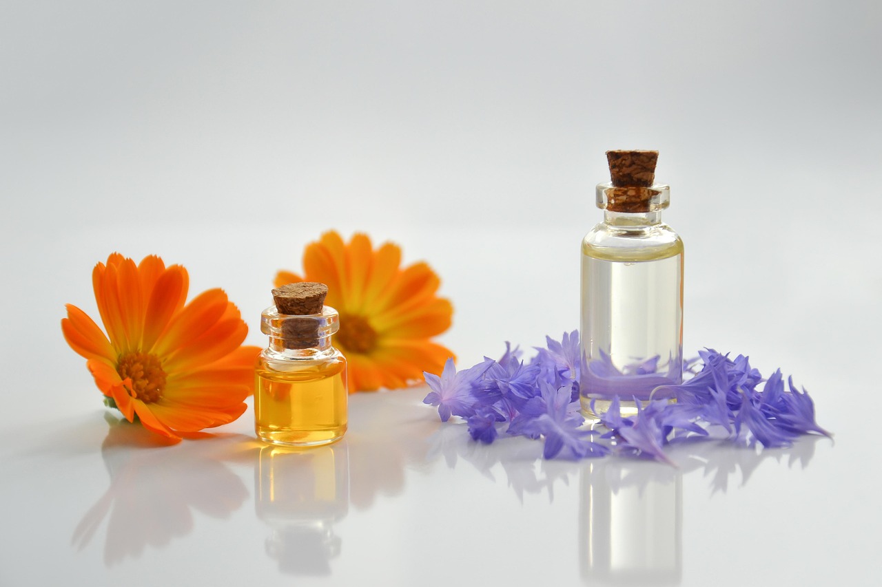 Dois frascos de vidro com rolha, líquidos amarelo e transparente, flores laranjas e lilases numa superfície branca