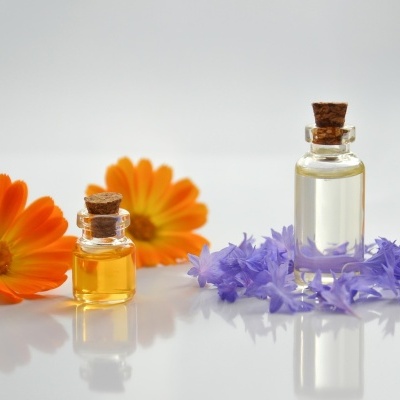 Dois frascos de vidro com rolha, líquidos amarelo e transparente, flores laranjas e lilases numa superfície branca