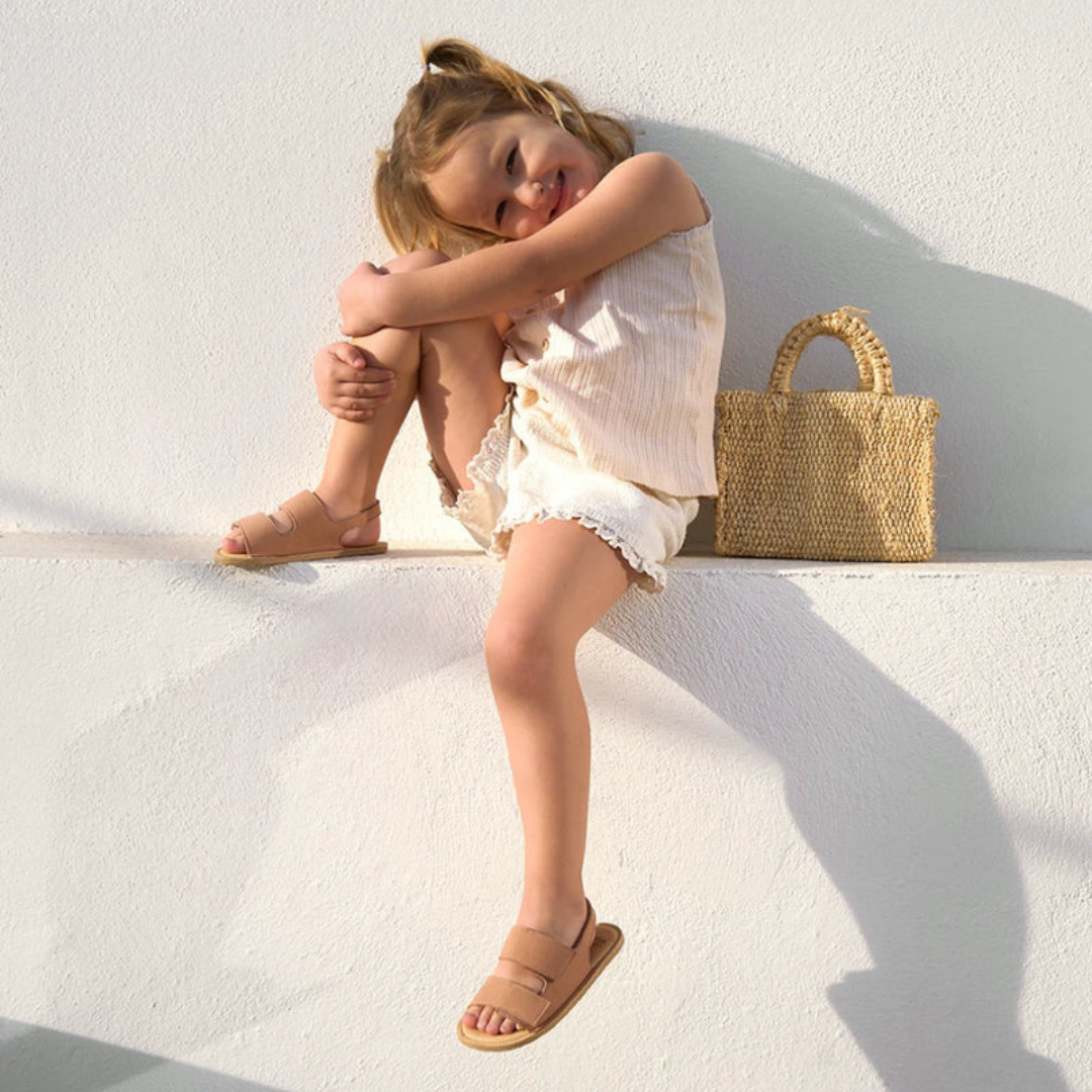 Criança com roupa clara e sandálias castanhas sentada ao lado de mala trançada natural