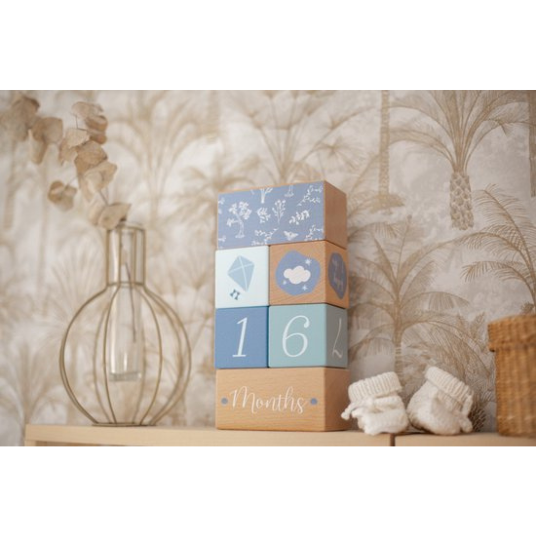 Blocos de madeira coloridos empilhados com número 16 e a palavra 'Months'. Ao lado, vaso de vidro e meias de bebé brancas.