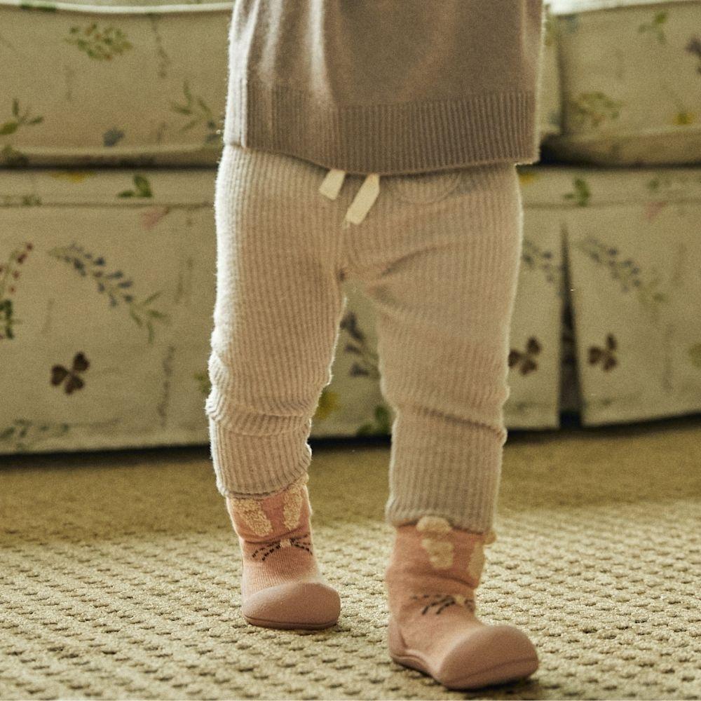 Criança com calças de malha bege e meias cor de rosa em tapete bege
