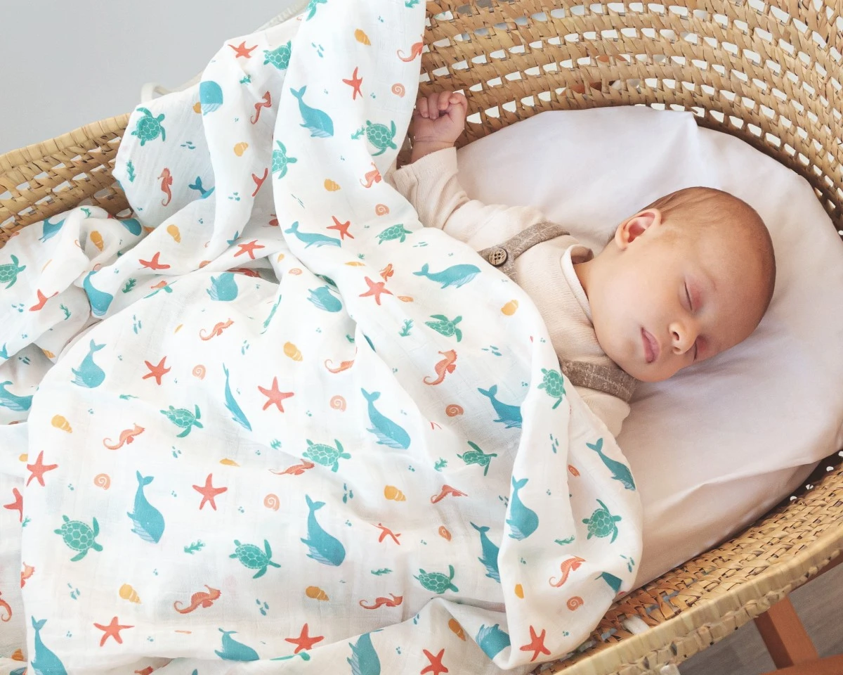 Manta de bebé branca com padrões de animais marinhos coloridos numa cesta de vime.
