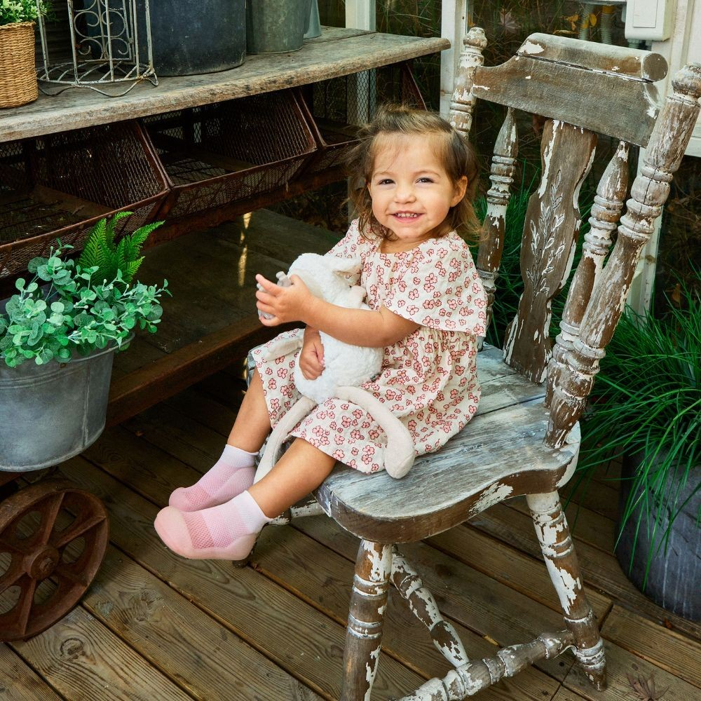 Menina sorridente sentada numa cadeira rústica com vestido floral e meias cor-de-rosa, segurando peluche branco