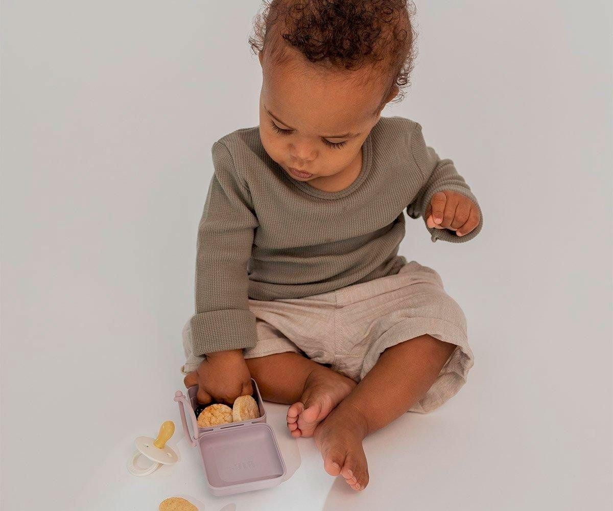 Bebé sentado visto de cima com roupa castanha e bege, recipiente lilás com bolachas e chupeta ao lado.