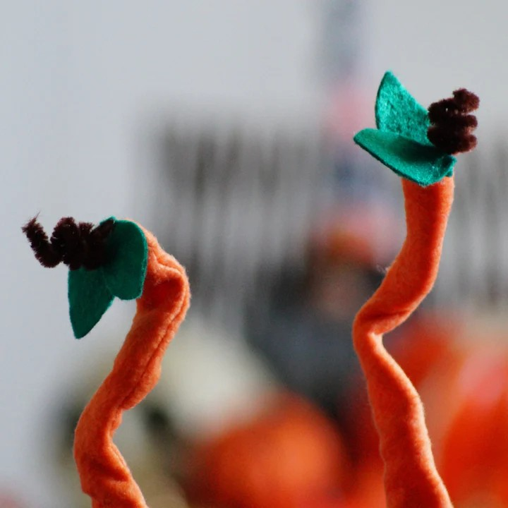 Dois objectos decorativos em forma de cenoura laranja com folhas verdes e pompons castanhos.