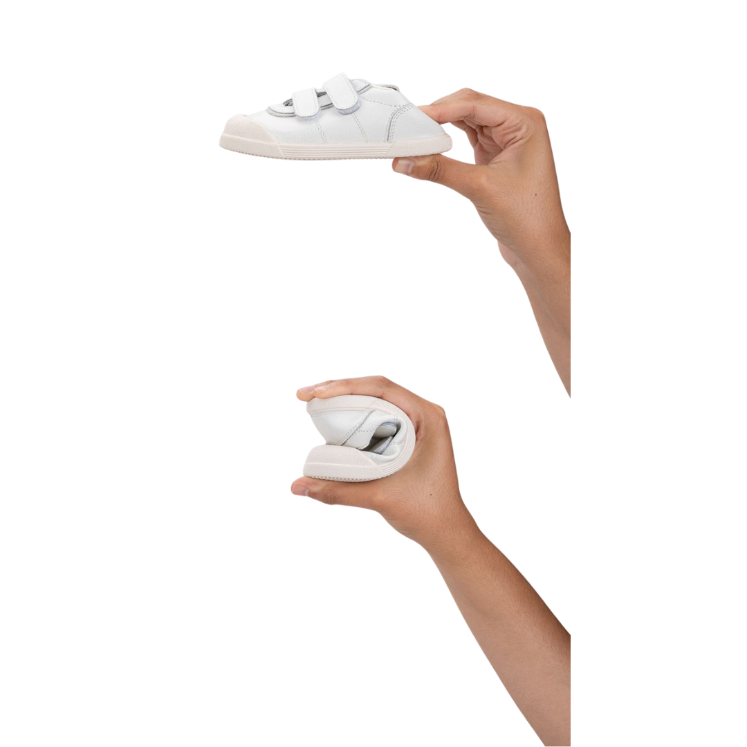 Tênis infantil branco com velcro e sola bege, dobrado e segurado por mãos.