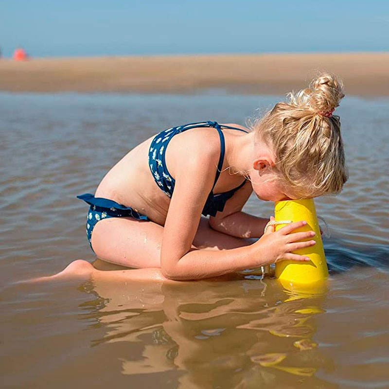 Criança de bikini azul com padrão a brincar na água rasa da praia segurando copo amarelo