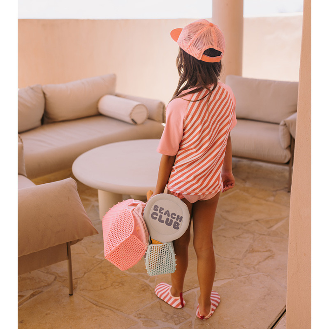 Criança vestida para praia com boné e t-shirt rosa com riscas, segurando raquetes de beach tennis e bolsa cor-de-rosa num ambiente com sofás beige.