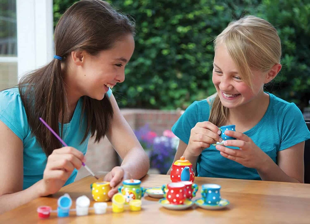 Conjunto de chá de brinquedo colorido com padrão de bolinhas em mesa de madeira, meninas pintando.