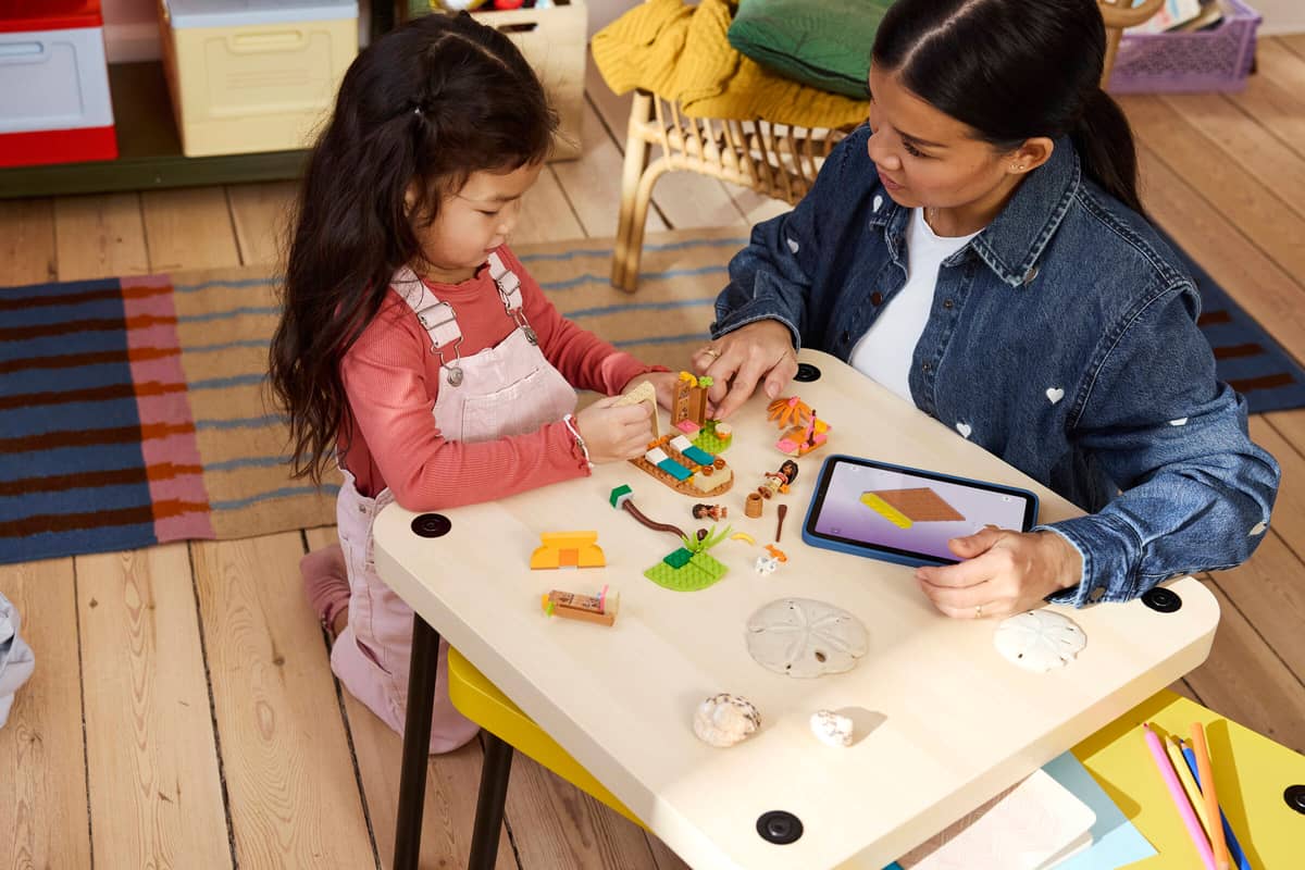 Criança e adulto brincando com peças de construção coloridas numa mesa branca com tablet, conchas, em ambiente interior com chão de madeira.